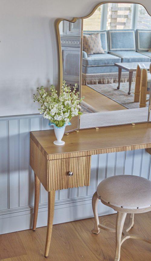 Wooden vanity table with a round stool, featuring a vase of white flowers. The table has a mirror reflecting part of a bed. The setting has light-colored walls and paneling.