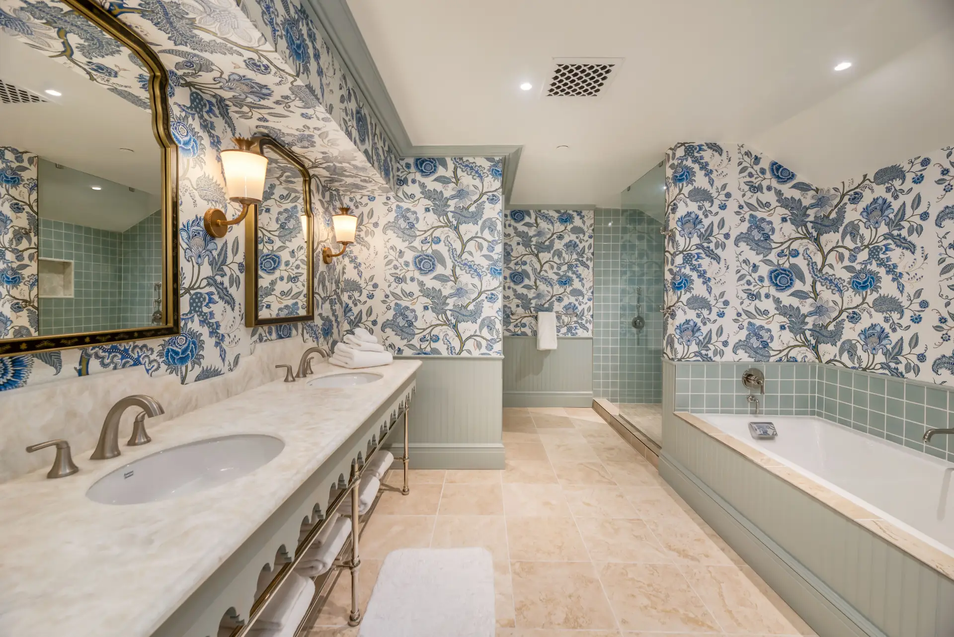 Spacious bathroom with golden accents, floral-patterned walls, and soft sea-green tones, featuring dual sinks for added comfort.